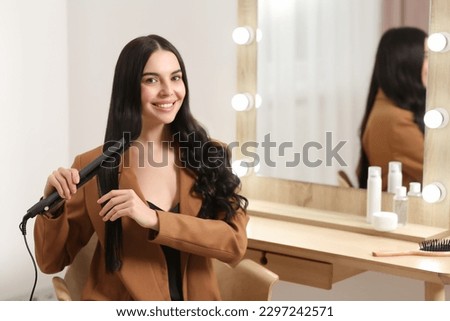Beautiful happy woman using hair iron near mirror in room. Space for text Royalty-Free Stock Photo #2297242571