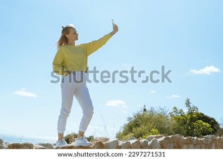 A girl of European appearance is standing in white pants and a yellow jacket taking a selfie overlooking the sea