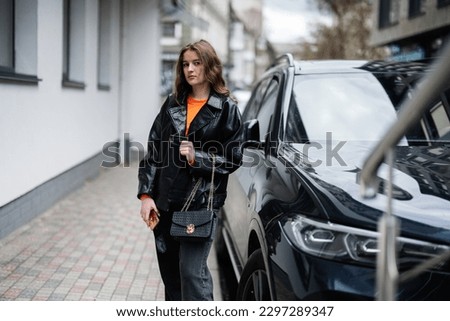Young girl in leather jacket posed in urban city, holding credit card with handbag and phone against black business car.