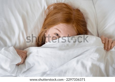Closeup of Lovely young Woman Teenage Girl Lies in Bed Covered with Blanket. Top view of Girl is Shy and Cover of her Face with Blanket. Female Smiling and Looking at Camera. Healthy Morning Routine Royalty-Free Stock Photo #2297336371