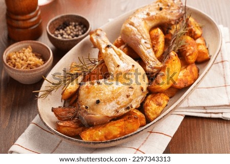two oven-roasted organic chicken thighs on roasted potato wedges spiced with chili and paprika on oval ceramic plate, rosemary and black pepper. Checkered kitchen towel, brown wooden tabletop Royalty-Free Stock Photo #2297343313