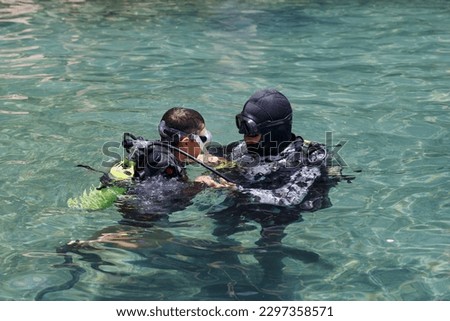 Diving with aqualung lesson, instructor giving scuba diving lessons to a kid. Trainer teaching a little boy to dive. Two scuba divers dressed in diving suit is preparing to dive in shallow sea. Royalty-Free Stock Photo #2297358571