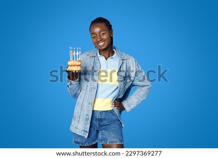 Cheery smiling cute young african american lady 20s wearing casual outfit and bright makeup posing with birthday cake delicious colorful donuts with lit candles, isolated on blue studio background Royalty-Free Stock Photo #2297369277