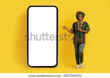 Happy smiling pretty young black lady in bright national african costume pointing at huge cell phone with white empty screen, isolated over yellow studio background, mockup, full length