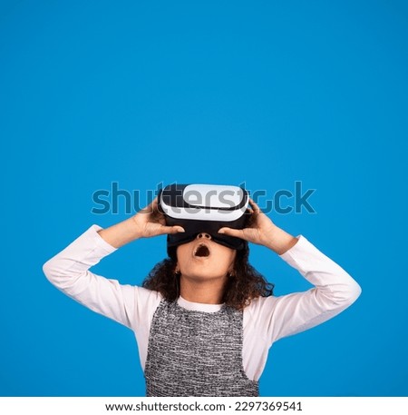 Shocked black little girl in VR glasses plays online game, look up with open mouth, has fun isolated on blue background, studio. Modern technology, childhood, emotions and offer