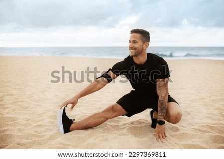Young strong sporty man in sports clothes doing warm up training, stretching legs on beach by seaside outdoors, looking aside and smiling, free space