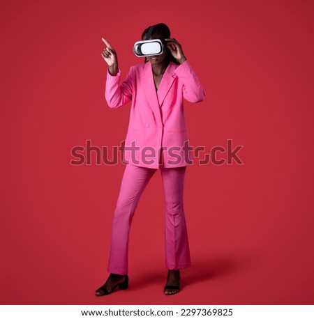 Black Woman Wearing Pink Suit And Vr Headset Enjoying Virtual Reality Experience, Full Length Of African American Female In Fashionable Clothes Touching Air With Finger, Standing On Red Background