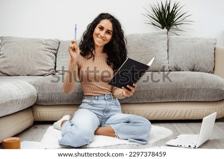 Young beautiful Arab woman holds a notebook in her hand and makes a wish list. She took a pen and is ready to paint everything point by point. The girl sits on the floor and smiles brightly at home. Royalty-Free Stock Photo #2297378459