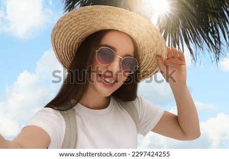 Smiling young woman in sunglasses and straw hat taking selfie under palm tree on sunny day