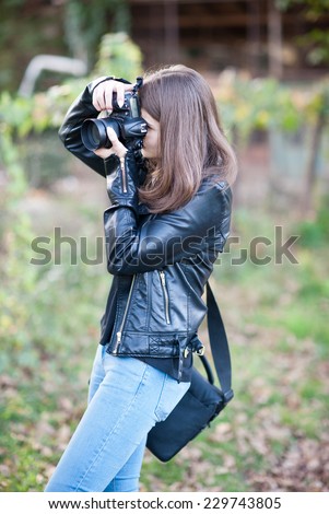 Attractive young girl taking pictures outdoors. Cute teenage girl in blue jeans and black leather jacket taking photos in autumnal park. Outdoor portrait of pretty teen having fun in park with camera