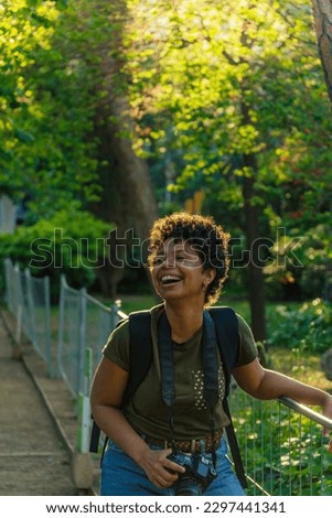 Happy young black African American woman photojournalist with dark curly hair laughing happily with open mouth looking at friend with photo camera hanging around neck in green park at sunset 