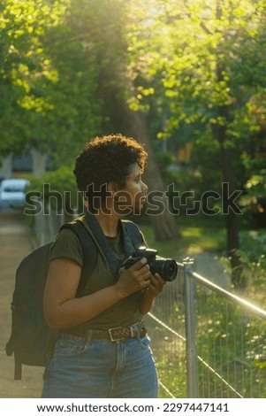 Young black African American female photojournalist with dark curly hair looking for something wonderful to photograph with photo camera in hand with the sun backlit behind
