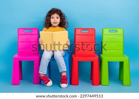 Full length cadre of young excited wavy hair schoolgirl reading her favorite fairy tail book sit chair isolated on blue color background