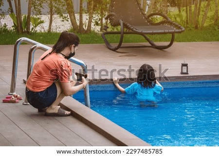 Asian mother wear face mask use smart phone to take picture her lovely daughter in blue swimming suit play with clear blue water. Happiness mom and daughter at pool. Happy family relationship outdoor.