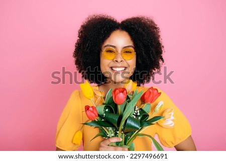Happy lovely brazilian or african american woman with curly hair, with sunglasses, in yellow dress, holds bouquet of colorful tulips, received gift, looks at camera, smile, isolated pink background