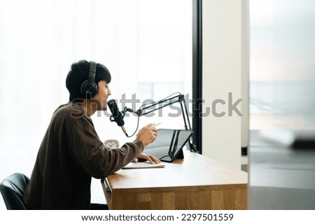 Young asian man host streaming podcast with condenser microphone work on tablet at small broadcast home studio. Content creator blogger recording voice over radio interview guest conversation Royalty-Free Stock Photo #2297501559