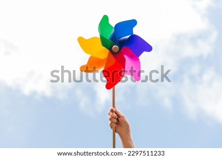 ecology, environment and sustainable energy concept - close up of hand holding multicolored pinwheel over blue sky Royalty-Free Stock Photo #2297511233