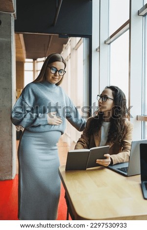 A pregnant woman is working in the office with a colleague on a new project, they are looking at a laptop. The work of female managers in a modern, spacious office