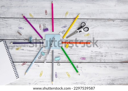 Bright, multicolored pencils for drawing in an album, colored paper clips, a blue notepad for notes, scissors, a pen, a metal sharpener and clips are folded neatly in a circle on wooden background.