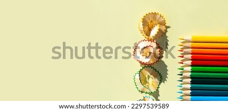 A large number of colored pencils with wooden shavings are sharpened and lie on a light yellow background with a clear shadow from natural light on a bright, sunny day.