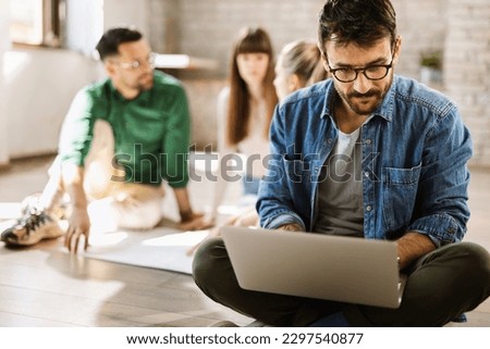 Young enterpreneur using computer while sitting on the floor in the office. There are people in the background Royalty-Free Stock Photo #2297540877