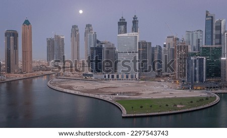 Cityscape of skyscrapers in Dubai Business Bay with peninsula on water canal aerial night to day transition with Moon setting down. Modern skyline with illuminated towers and waterfront before sunrise