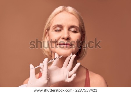 Portrait of a beautiful blonde middle-aged woman with closed eyes getting an injection in the chin on a beige background. Hands in white gloves hold the chin and syringe.Medical procedure.Face lift. Royalty-Free Stock Photo #2297559421