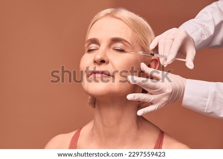Smoothing out wrinkles.Portrait of an older adult woman with closed eyes doing a beauty injection. Hands in white gloves hold a syringe.Face lift. Royalty-Free Stock Photo #2297559423