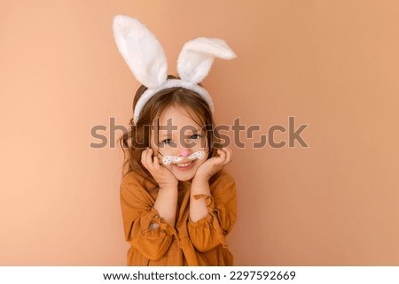 A sweet, pleasant little girl dressed up in an Easter bunny costume looks in surprise at the camera with big blue eyes, pressing the boats to her cheeks.
