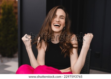Beautiful young woman happy and excited expressing winning gesture. Successful and celebrating victory, triumphant. Woman sitting at home outdoors, wear pink pants, black tank top and accessories.