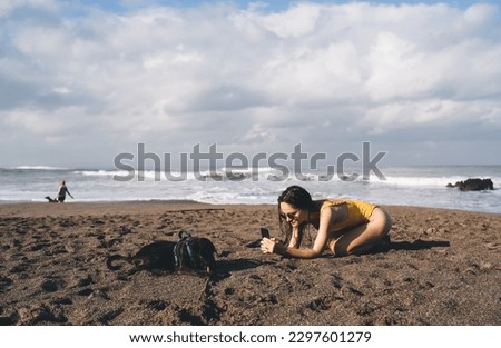 Side view of young female in sunglasses kneeling on sandy beach near pet dog and taking picture while passing time against waving sea and cloudy blue sky