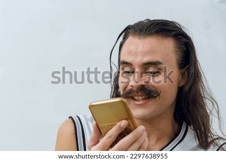Latino young person of non-binary gender and Argentinian ethnicity, with long hair and mustache, is smiling and using the phone in his room, copy space.