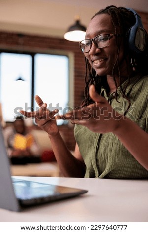 Portrait of student with wireless headphones talking casual in video call conference with friends while boyfriend is relaxing. African american freelancer working remote having online meeting.