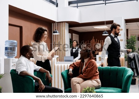 Woman welcoming hotel guests in lobby, providing excellent luxury service to people at resort. Young manager discussing about staff and accommodation with tourists, giving advice. Royalty-Free Stock Photo #2297640865