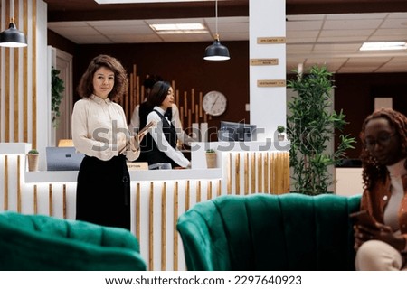 Portrait of hotel manager with records standing in reception desk lobby area to check concierge service as supervisor. Female lodging manager and administrator welcoming guests. Royalty-Free Stock Photo #2297640923