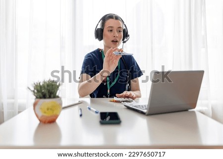 Young beautiful female doctor wearing headphones, making online webcam video call on laptop screen and gives advice to patient. Videocall view. Tele medical concept of health care Royalty-Free Stock Photo #2297650717