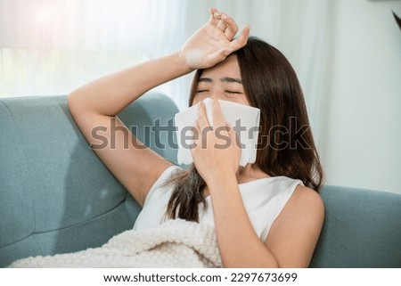 Sick woman sitting under blanket on sofa and sneeze with tissue paper in living room, Asian young female blowing nose sneezing in tissue at home, fever caught cold Royalty-Free Stock Photo #2297673699