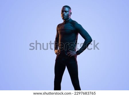 Serious African American male athlete in black sportive turtleneck and leggings standing on blue backdrop with hands on waist and looking at camera Royalty-Free Stock Photo #2297683765