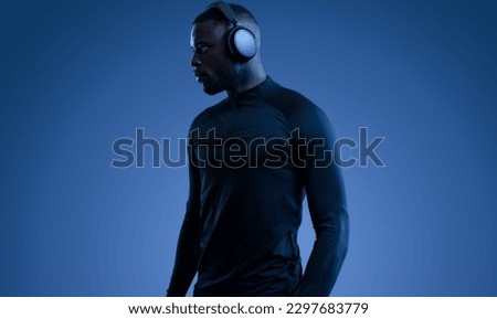 Confident young African American sportsman in black activewear listening to music in modern wireless headphones and looking away against blue background Royalty-Free Stock Photo #2297683779