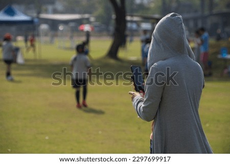 Mom standing and taking pictures of her daughter playing football in a school tournament on a sideline with a sunny day. Sport, outdoor active, happy family and soccer mom and soccer dad concept.