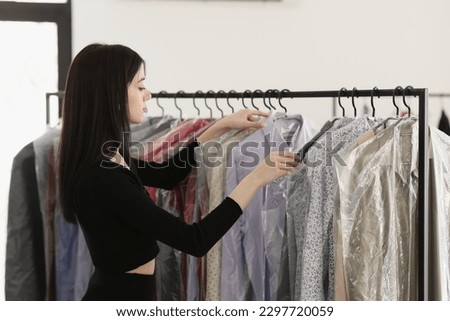Brunette dry cleaning worker looks for client coat on rack. Young woman goes over dry-cleaned clothes on hangers under film to find right item