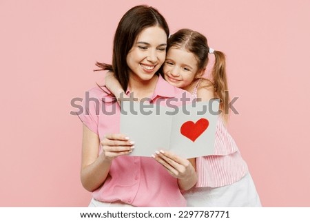 Happy smiling woman wear casual clothes with child kid girl 6-7 years old. Daughter give mother postcard with heart, hug and cuddle isolated on plain pastel pink background. Family parent day concept