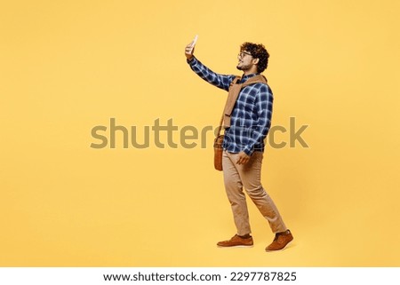 Full body young teen Indian boy IT student wear casual clothes shirt glasses bag doing selfie shot on mobile cell phone isolated on plain yellow color background High school university college concept