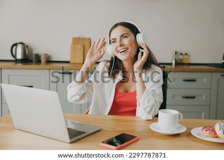 Young smiling housewife IT woman wearing casual clothes headphones hold use working on laptop pc computer listen music eat breakfast cooking food in light kitchen at home alone. Healthy diet concept