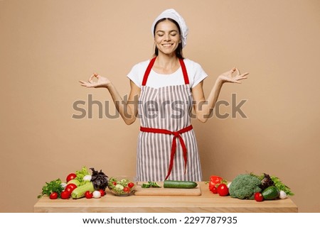 Young calm housewife housekeeper chef cook latin woman in apron toque chefs hat work at table kitchenware do yoga om aum gesture isolated on plain pastel light beige background. Process cooking food