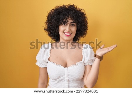 Young brunette woman with curly hair standing over yellow background smiling cheerful presenting and pointing with palm of hand looking at the camera. 