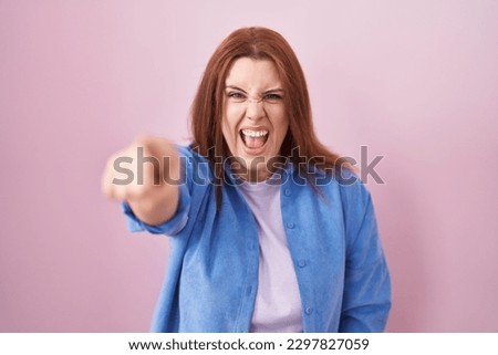 Young hispanic woman with red hair standing over pink background pointing displeased and frustrated to the camera, angry and furious with you  Royalty-Free Stock Photo #2297827059