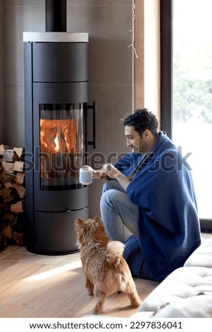 Young man sitting close to the fireplace with his cute joyful dog and warming in cold winter day	