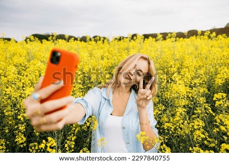 Beautiful young blonde cheerful woman, taking a selfie with a red phone, amidst a field of blooming yellow rapeseed flowers