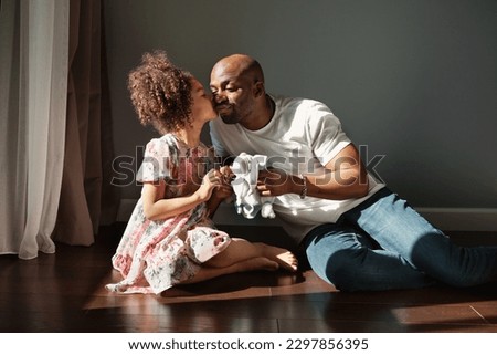 Happy African American father and his daughter playing toy together. Baby girl kissing dad sitting on floor indoors. Lifestyle portrait, sunny.